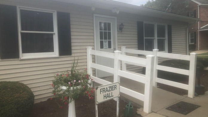 Exterior single-story light-sided building with windows, entrance door, and Frazier Hall identification sign