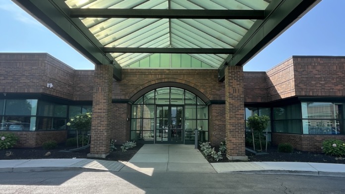 Exterior entryway with modern glass canopy, brick building facade, landscaping, and concrete walkway approach