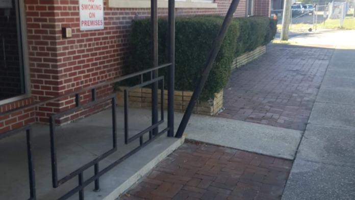 Exterior entrance area with brick building facade, covered awning, metal railings, and paved walkway leading to facility entrance