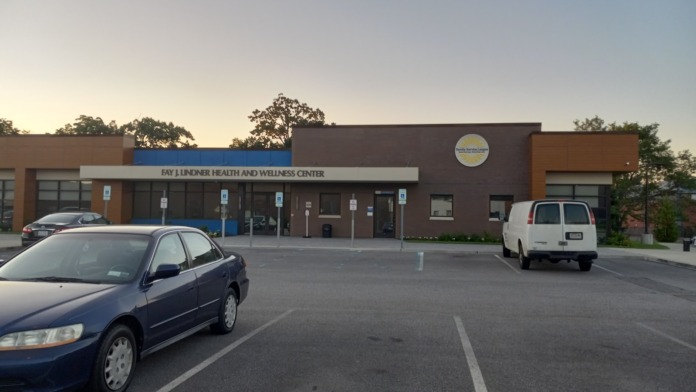 Exterior view of treatment facility showing main entrance with signage, parking lot with vehicles, and brown brick facade