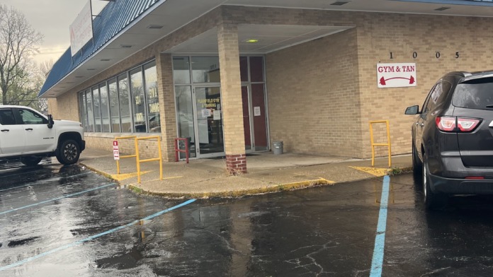 Exterior view of brick building with blue metal roof, main entrance, and parking area with vehicles