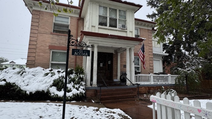 Exterior front view of brick building with snow, white picket fence, sign post, covered porch, and landscaping