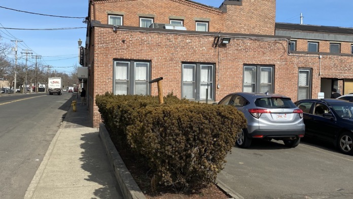 Exterior front of brick facility showing main entrance doors, visitor parking spaces, and landscaped evergreen shrubs