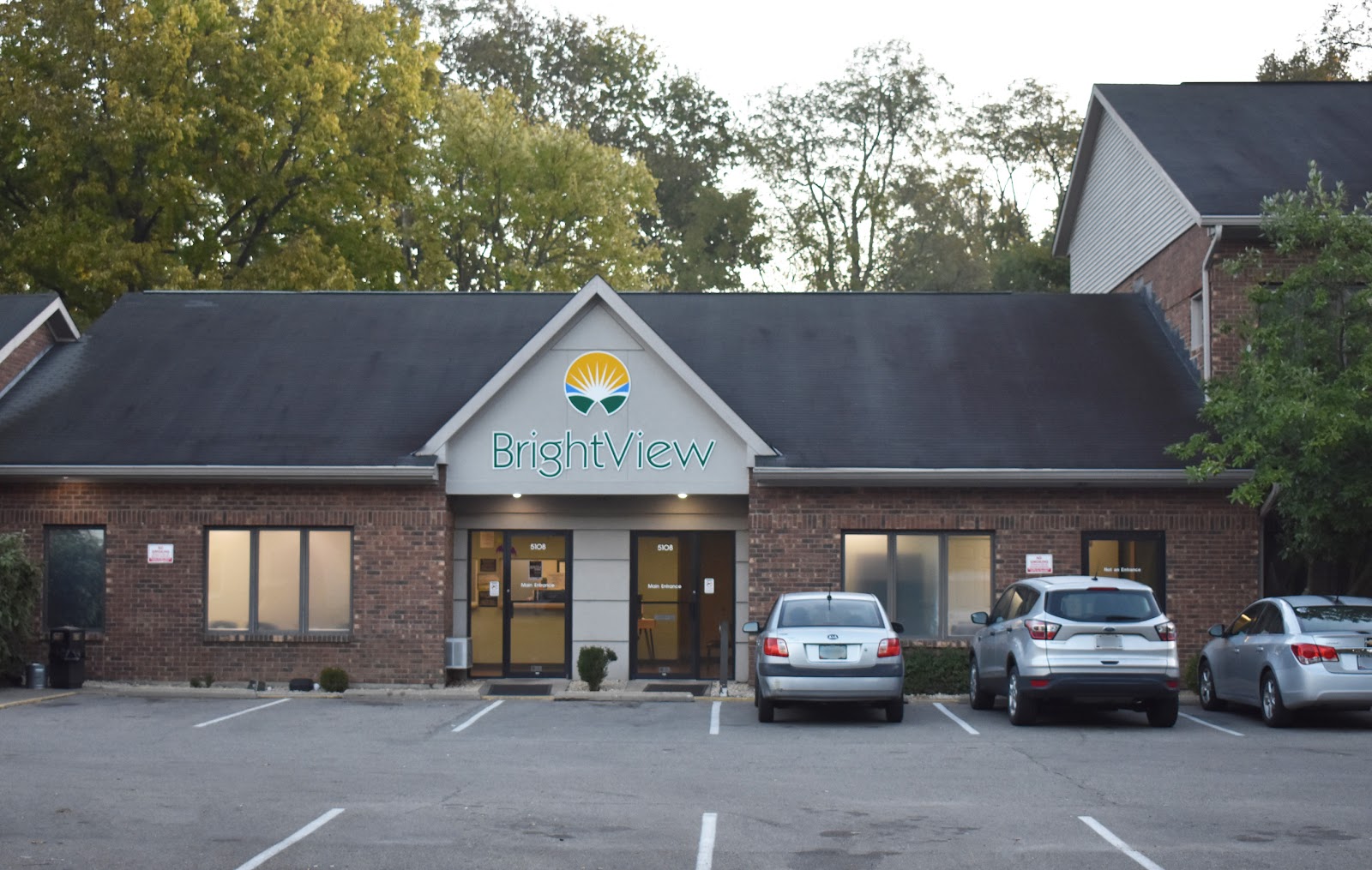 Exterior front view of brick facility with BrightView signage, gabled roof, windows, and parked vehicles at dusk.