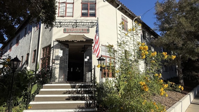 Exterior front-facing view showing two-story white building with entry steps, windows, flag, and flowering planters.