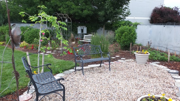 Outdoor garden area with metal benches, climbing trellis, flowering plants, ornamental grasses, white fencing, and facility building