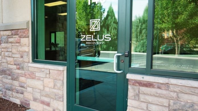 Exterior entrance with green-framed glass doors, stone building base, decorative shrubs and greenery, modern architectural details