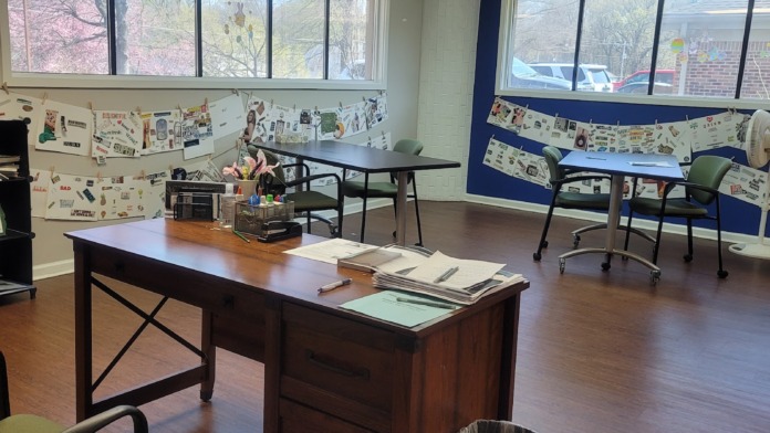 Interior activity room with large windows, tables, chairs, bulletin boards with posted materials, and wooded views