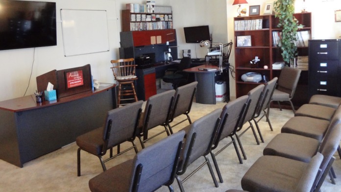 Interior room with multiple padded chairs arranged in rows, desks, bookshelves, monitor, and office furnishings