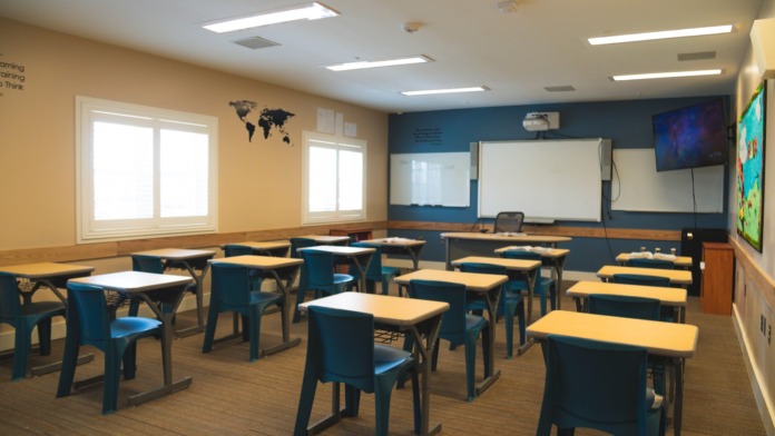 Interior classroom with student desks, chairs, whiteboards, windows, world map, and educational materials