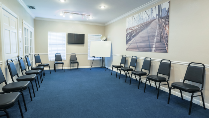 Interior meeting space with blue carpet, black stacking chairs arranged in semi-circle formation, whiteboard, TV, and landscape wall art.