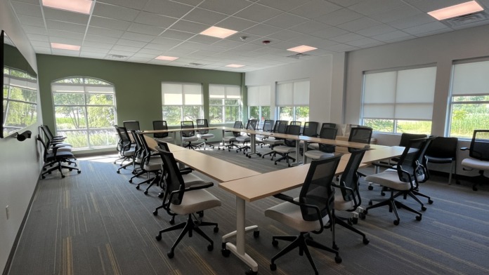Interior conference space with multiple tables, black office chairs, large windows with natural light, and whiteboard