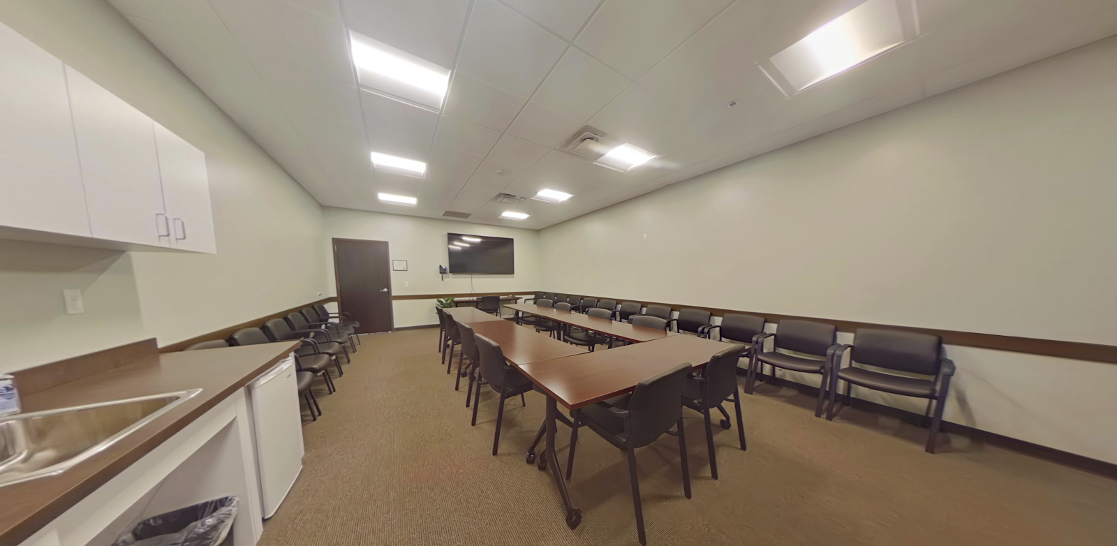 Interior meeting room with long wooden table and multiple black chairs arranged in semi-circle.