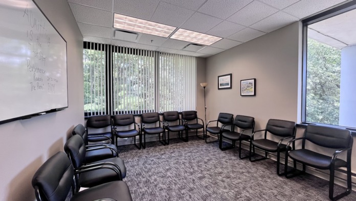 Interior therapy room with multiple black chairs, whiteboard, framed artwork, and large windows with vertical blinds