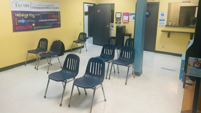 Interior waiting or group room with yellow walls, blue support column, multiple black chairs, and facility signage.
