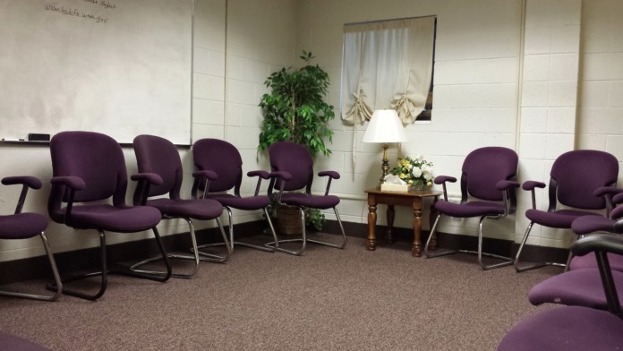 Interior group session room with multiple purple cushioned chairs arranged in circle, plants, and wall mirror