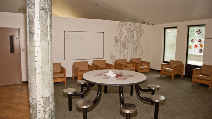Interior therapy space with white walls, decorative birch tree mural, round table with attached seating, and wood chairs arranged in circle