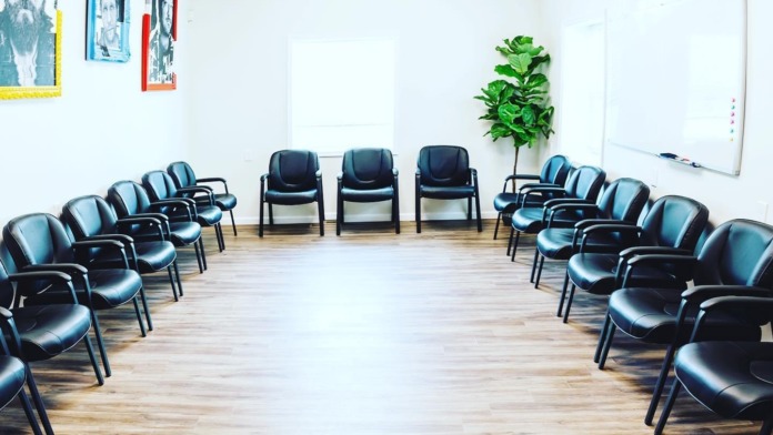 Interior group therapy room with multiple black chairs arranged in semi-circle configuration, whiteboard, and colorful artwork on walls