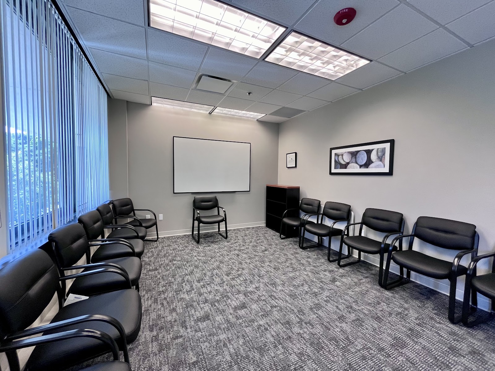 Interior therapy room with multiple black chairs arranged in rows, projection screen, and window with blinds