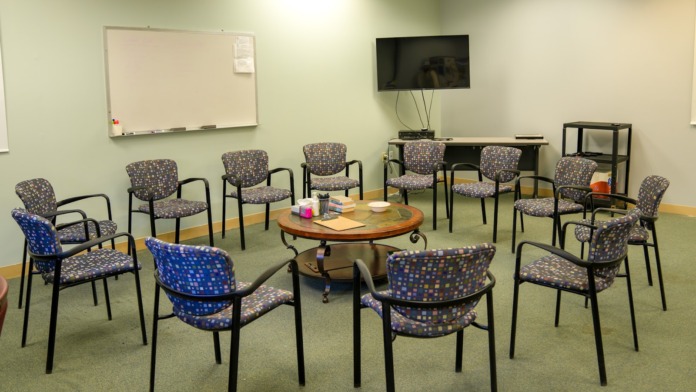 Interior therapy room with multiple patterned chairs arranged in circle around central table, whiteboard, and wall-mounted television