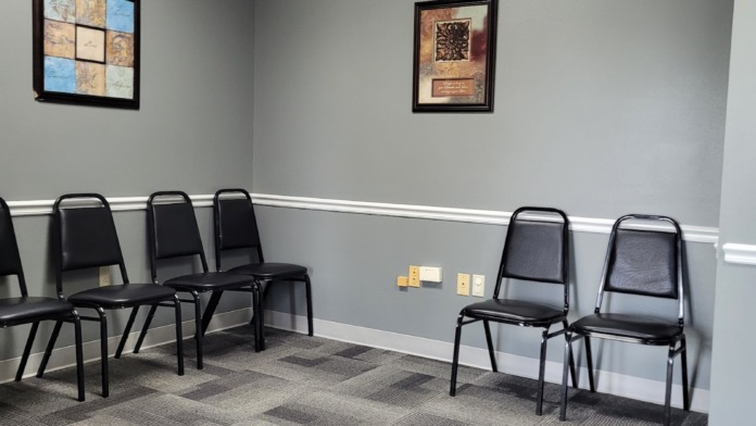 Interior waiting room with black stackable chairs, framed wall art, patterned carpet flooring, and drop ceiling lighting