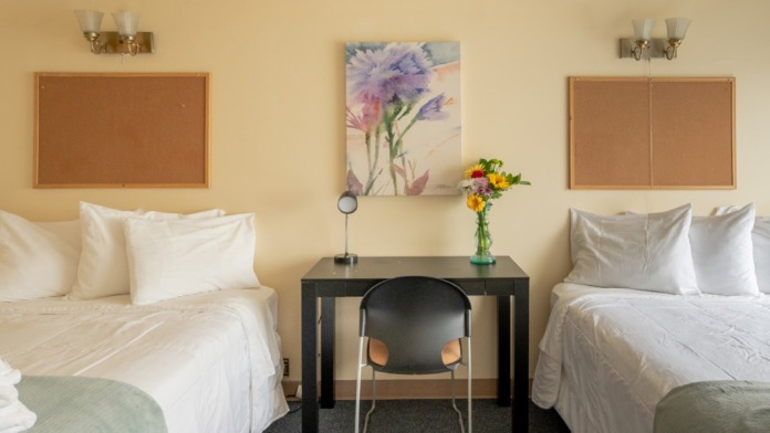 Interior bedroom with two twin beds, pale yellow walls, cork bulletin boards, central side table with flowers, and dark flooring