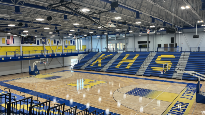 Interior gymnasium featuring basketball court, blue and yellow seating bleachers with school letter, hardwood floor, and overhead lighting