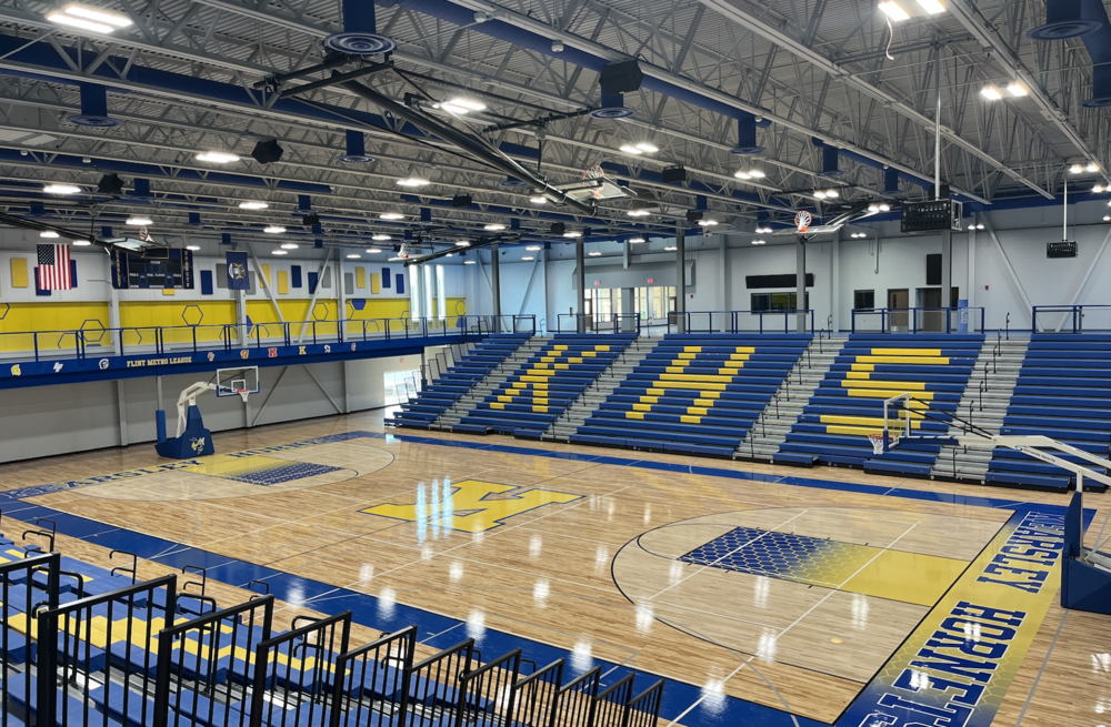 Interior gymnasium featuring basketball court, blue and yellow seating bleachers with school letter, hardwood floor, and overhead lighting