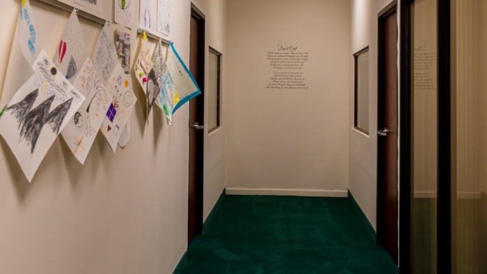 Interior hallway corridor with patient artwork and creative displays on walls, green flooring, doors to patient rooms, and ceiling lights