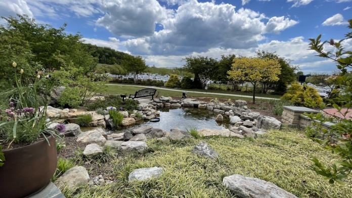 Outdoor therapeutic garden with koi pond, rock landscaping, flowering plants, benches, walking paths, and facility grounds backdrop