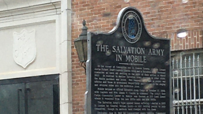 Exterior of brick building with white-trimmed entrance, white picket fence, and historical marker plaque for The Salvation Army in Mobile.