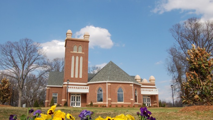 Exterior view of red brick church with bell tower, arched stained glass windows, and colorful flower beds