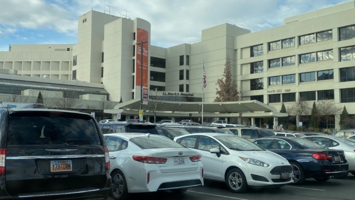 Exterior view of hospital complex showing multiple buildings, parking lot, red banner signage, and adjacent structures