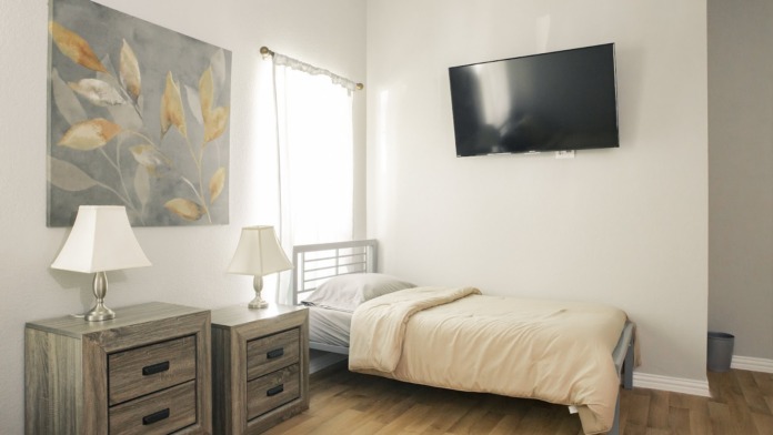 Interior patient bedroom with single bed, nightstand, lamp, artwork, and window with natural light