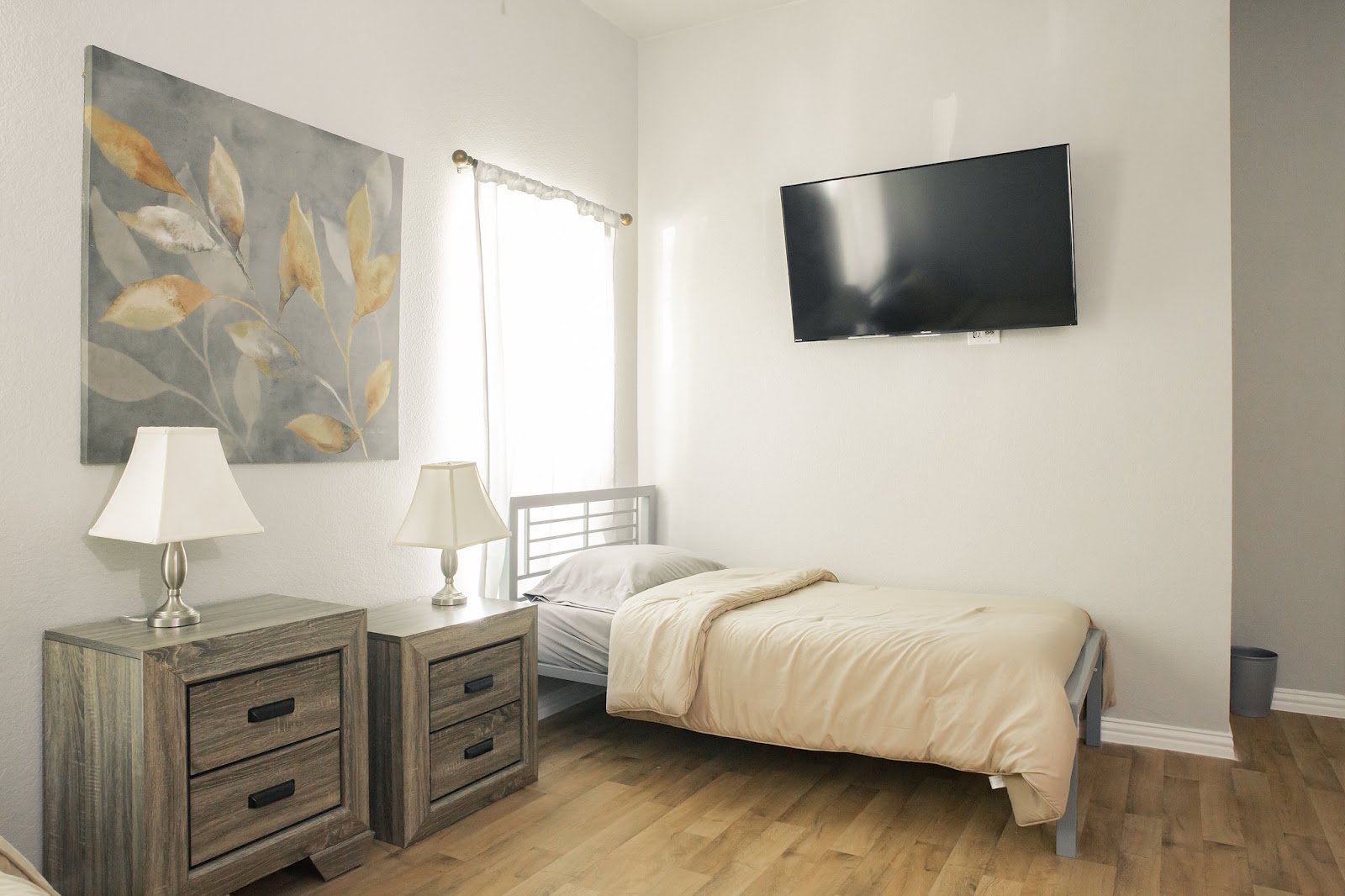 Interior patient bedroom with single bed, nightstand, lamp, artwork, and window with natural light