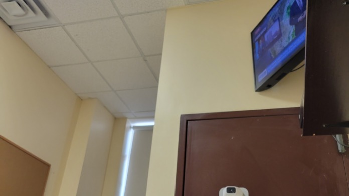 Interior hallway with mounted television screen, decorative plants, drop ceiling, and informational yellow notice