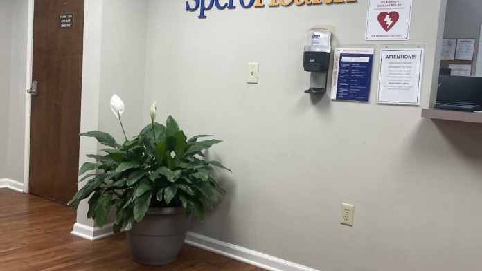 Interior hallway with SperoHealth signage, plant decoration, AED equipment, white walls, wooden flooring, and door entry