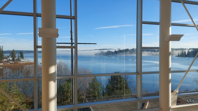 Interior view through large windows showing calm blue water, forested shoreline, and natural daylight