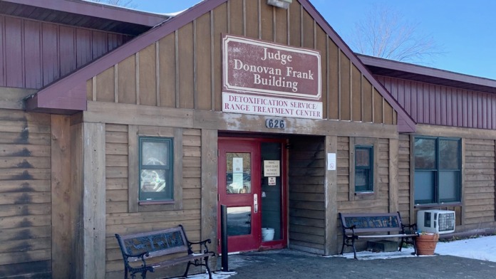 Exterior of detoxification service building with wooden siding, red entrance door, benches, winter setting, and facility signage