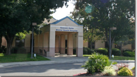 Exterior view of single-story hospital building with blue roof accent, main entrance doors, landscaped grounds with shrubs and mature trees