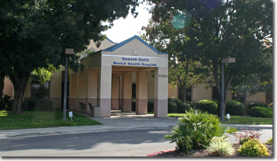 Exterior view of single-story hospital building with blue roof accent, main entrance doors, landscaped grounds with shrubs and mature trees