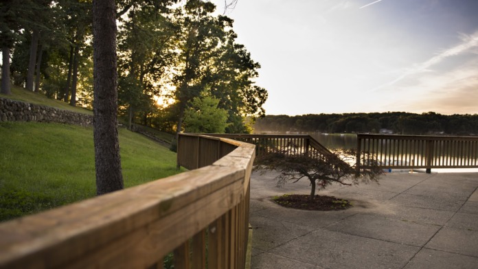Outdoor deck area with wooden railings, green lawn, mature trees, and calm lake water view