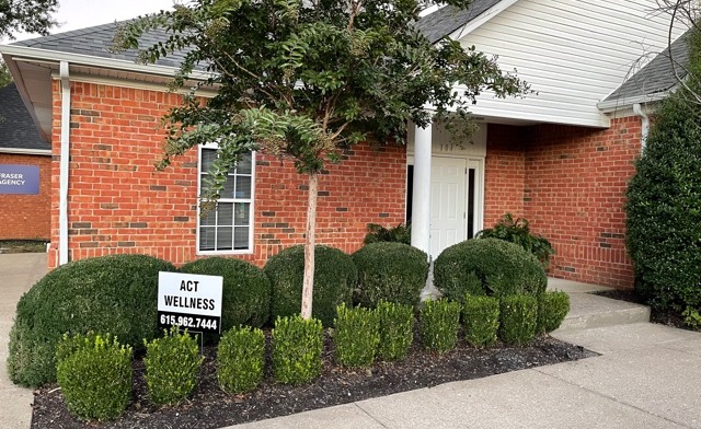Exterior view showing manicured shrubs, landscaping, brick building side, and paved walkway area