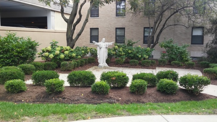 Exterior garden area with religious statue centerpiece, flowering plants, mature trees, brick building backdrop, and manicured landscaping