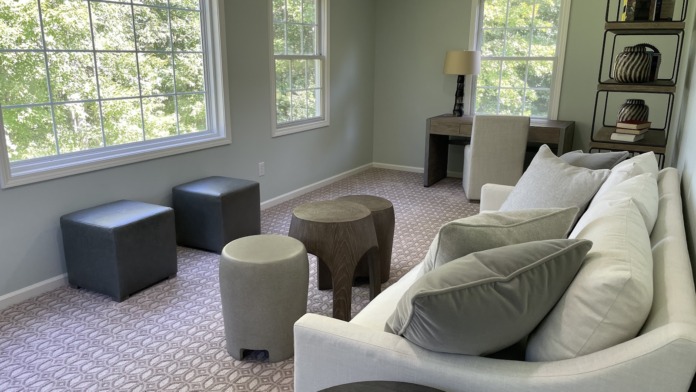 Interior lounge with cream sofa, dark ottoman seating, wooden side tables with lamps, and large windows with natural light