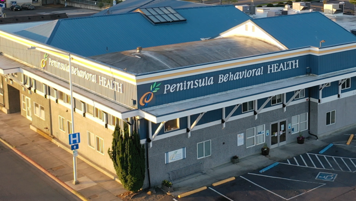 Exterior view of Peninsula Behavioral Health facility showing multi-story structure with blue signage, entrance doors, windows, and parking lot from elevated angle