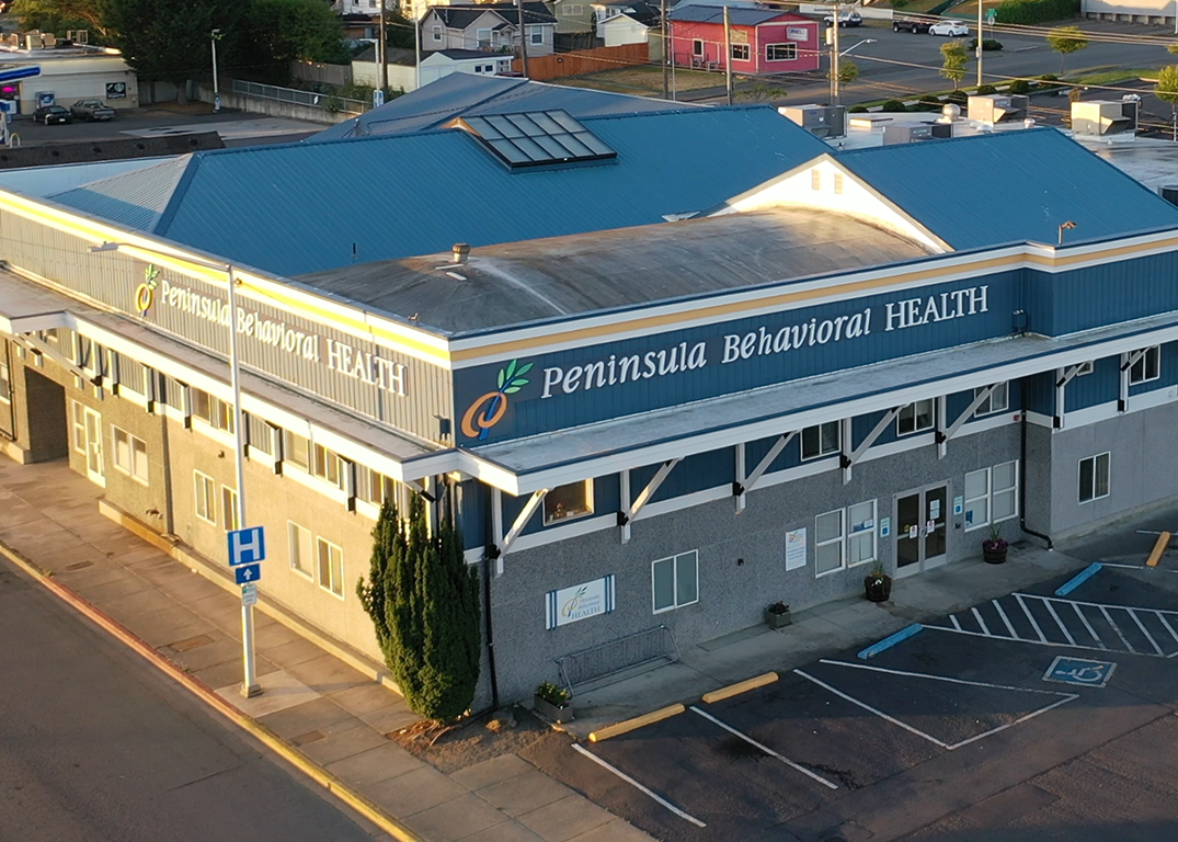 Exterior view of Peninsula Behavioral Health facility showing multi-story structure with blue signage, entrance doors, windows, and parking lot from elevated angle