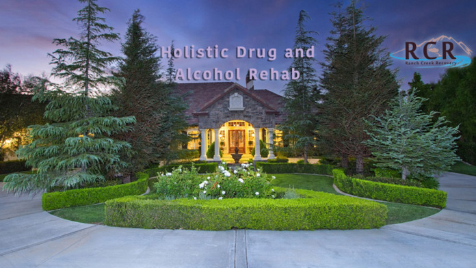 Exterior evening shot of stone treatment facility building with illuminated entrance, landscaped grounds, hedges, and text overlay describing holistic rehab services