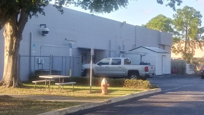 Exterior view of facility building with large white facade, parking area, picnic table, and mature trees
