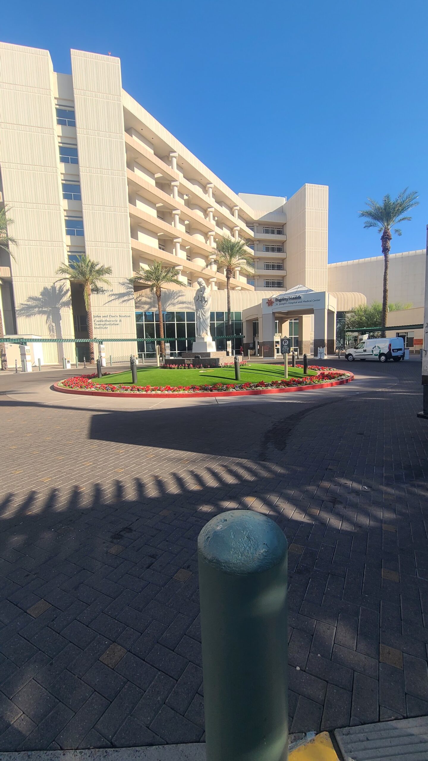 Exterior main entrance with palm trees, decorative flowering beds, modern facility signage, and paved courtyard area